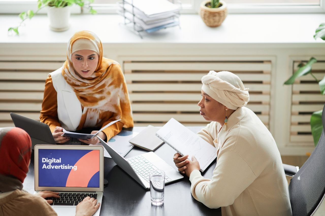 services-03 Three women in hijabs collaborating on digital advertising strategies around a table with laptops.
